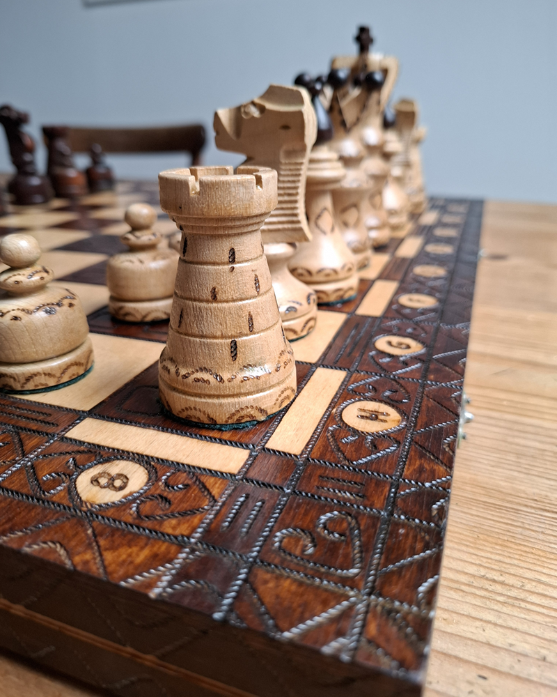 Hölzernes Schachspiel, fast auf Augenhöhe fotografiert. Man sieht einen Rand mit dunkelbraunem Holz und hellen Markierungen. Im Vordergrund sind einige helle, geschnitzte Schachfiguren zu sehen mit schönen Details. Im Hintergrund werden die Figuren unscharf, links hinten sieht man einige dunkelbraune Figuren. Das Schachspiel steht auf einem Holztisch, wie man am rechten Rand des Fotos erkennen kann.