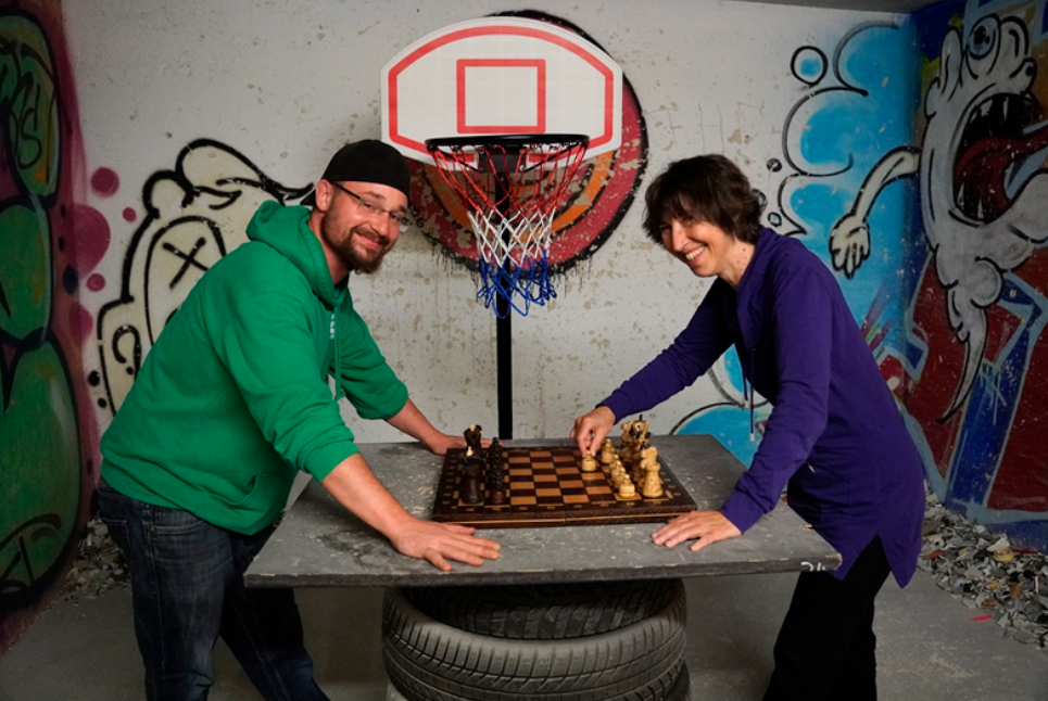 Roman Giesswein und ich stehen gegenüber über eine Tischplatte gebeugt, die auf einem Reifenstapel steht. In der Mitte steht das hölzerne Schachspiel, ich habe die Hand an einem Bauern, der ein Feld nach vorne gezogen ist. Im Hintergrund, zwischen uns, ist ein Basketballkorb zu sehen. Die Wand dahinter ist schmutzig weiß und teilweise sieht man Graffiti.