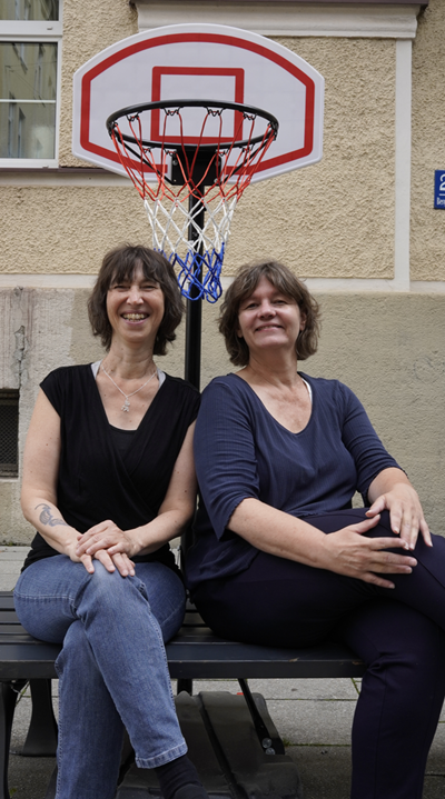Auf einer Bank, die an einem Bürgersteig steht, sitzen zwei mittelalterliche Frauen mit übereinander geschlagenen Beinen und lachen in die Kamera. Dahinter, zwischen und über ihren Köpfen ist ein Baskettballkorb zu sehen, der ein rot-weiß-blaues Netz hat. Dahinter eine Hausfassade.