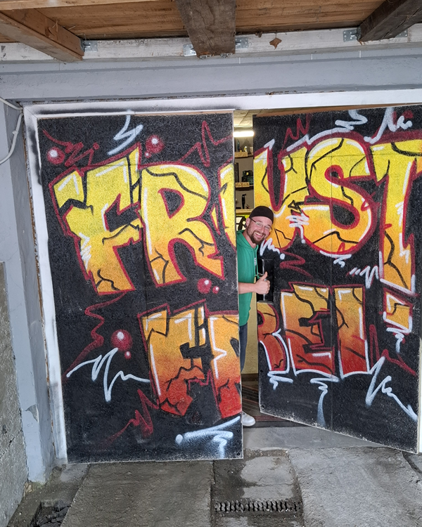 Die Eingangstüre des Unternehmens "Frust frei Landshut" hat den Schriftzug Frustfrei auf einem Metalltor aufgesprüht, farbenfroh von einem Graffitikünstler. Das Tor steht einen Spalt auf und der Eigentümer, Roman Giesswein, schaut aus dem Türspalt, lächelt freundlich und zeigt ein Daumen hoch.