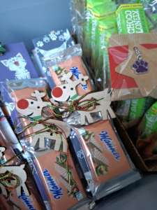 In Cellophan verpackte Mannerkekse. Weihnachtlich verpackt auf bezogenen Kartons mit Tiermotiven, darauf ein hölzernes Rentier und rot-grün-goldene Schnüre mit Schleife, daneben Schokolade mit einem glitzernden Pfotenanhänger.