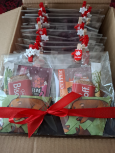 Jeweils zwei aus Papier gefaltete tassenförmige Taschen, in denen jeweils steckt: ein Teebeutel, ein Keks, Zucker, das ganze in Cellophan verpackt, mit einer roten Schleife umwickelt und kleine winterliche Holzklammern zur Deko. Man sieht einen Karton davon.