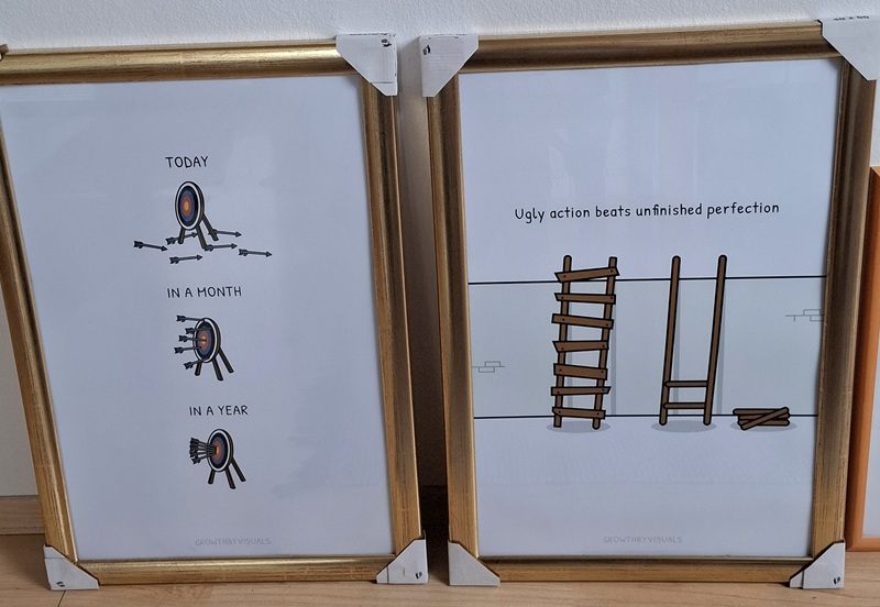 Zwei Poster mit Illustrationen in einem goldfarbenen Rahmen stehen nebeneinander. Das linke Poster zeigt drei untereinander stehende Zielscheiben. Die oberste hat die Überschrift „Today“ und zeigt lauter Pfeile, die neben der Scheibe liegen, weil sie nicht getroffen wurden. Darunter steht „In a month“. Auf dieser Zielscheibe sind mehrere Pfeile, die in der Scheibe stecken, aber verstreut auf verschiedenen Stellen. Die unterste Zielscheibe ist überschrieben mit „In a year“. In dieser stecken alle Pfeile in der Mitte. Das rechte Poster hat die Überschrift „Ugly action beats unfinished perfection“ und zeigt eine Mauer, an der zwei gezeichnete Leitern lehnen. Die linke ist vollständig, aber mit grobem Holz krumm und schief zusammengebaut. Die rechte ist perfekt gerade gebaut mit abgerundetem Holz, allerdings sind nur zwei Sprossen an der Leiter, die anderen Sprossen liegen auf einem Haufen rechts daneben. Auf beiden Postern steht unten im Bild in hellgrauer Farbe „growthbyvisuals“